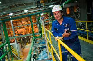 Man dressed in Cameco coveralls and hard hat leaning on a railing with pipes in background