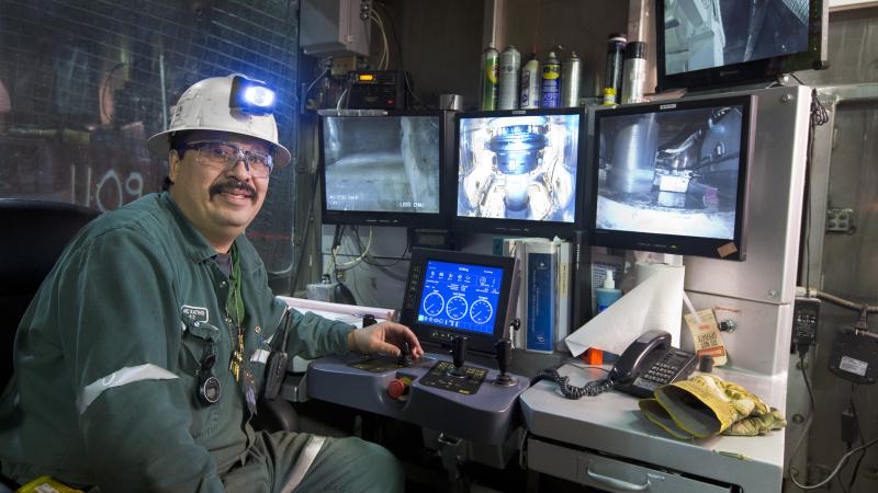 Man with moustache wearing hardhat in front monitors smiling at camera